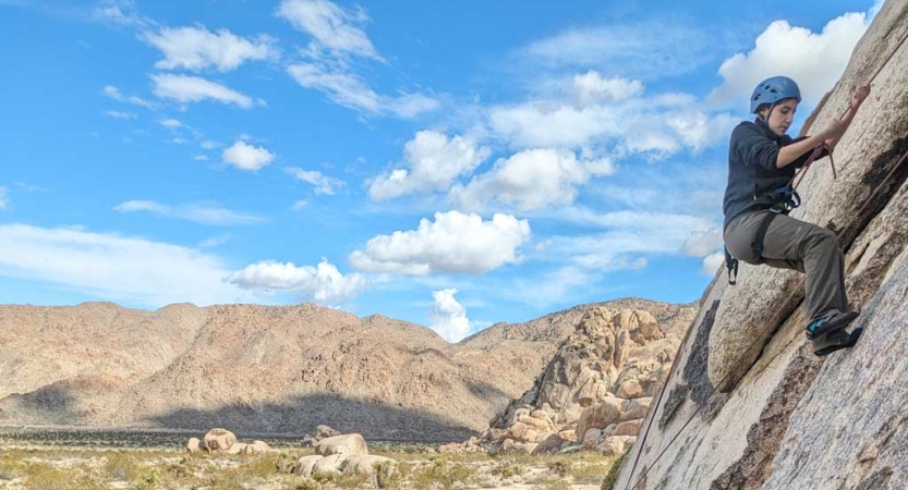 A person wearing safety gear is secured by ropes as they climb a rock wall. Behind them is a mountain range. 